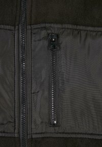 Svart fleece-fôret jakke med ytre nylonstoff. Har en sentral glidelås og en sidelomme med glidelås. Glatt tekstur, praktisk design.