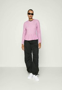 Růžový žebrovaný svetr s peplum spodním lemem, zkombinovaný s volnými černými cargo kalhotami a bílými teniskami. Model nosí tmavé sluneční brýle.