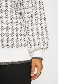 Main tenant deux bagues en or, reposant à côté d'un pull en tricot blanc à motifs avec des détails noirs et argent métallique, et d'un pantalon noir.