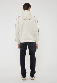 Felpa con cappuccio color beige chiaro, con strisce nere ai lati, in cotone, con design aderente. Abbinata a pantaloni scuri e sneakers bianche.