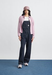 Νεαρή γυναίκα με ροζ καπέλο τύπου κάπα, ροζ πουκάμισο με κουμπιά πάνω από λευκή μπλούζα, σκούρα τζιν σαλοπέτα και λευκά αθλητικά παπούτσια, στέκεται σε μπλε χαλί.