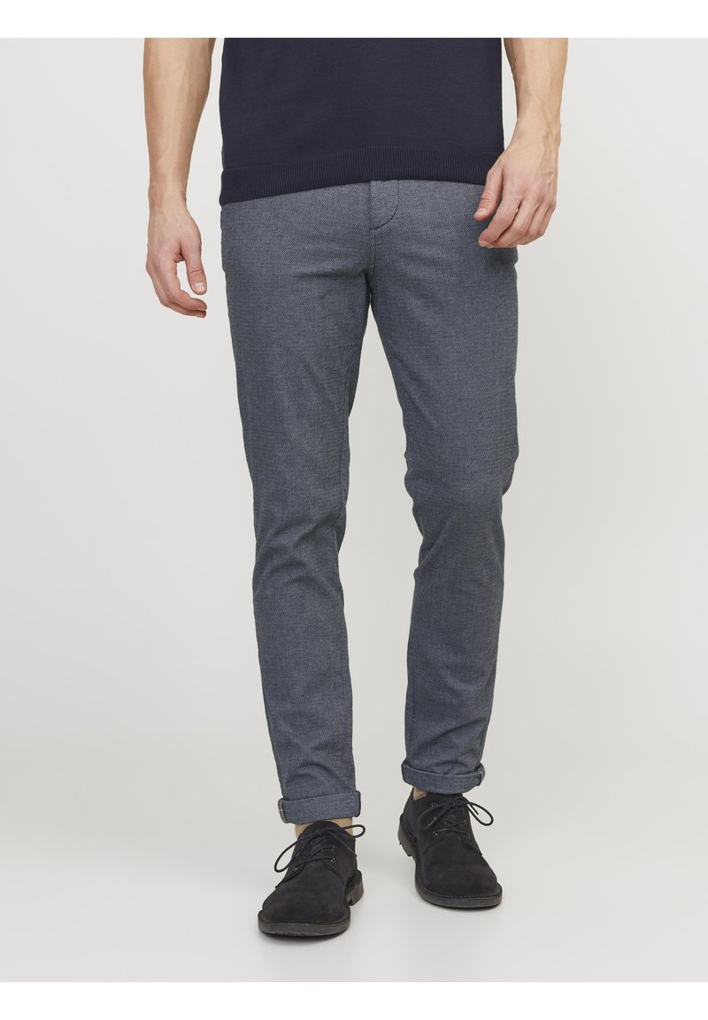 Homme portant un pantalon gris slim texturé roulé aux chevilles, avec des chaussures noires à lacets et un pull bleu marine foncé.
