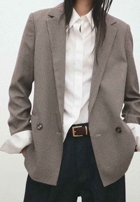 Bruin-witte houndstooth blazer met dubbelgeknoopte knopen, puntige revers en een ontspannen pasvorm, gecombineerd met een wit overhemd en donkere broek.