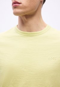 Κοντινό πλάνο ενός ατόμου που φοράει ανοιχτοκίτρινη μπλούζα με στρογγυλή λαιμόκοψη και μικρό λογότυπο "BOSS" στο στήθος.