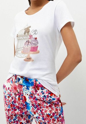 Weißes T-Shirt mit einem Grafikdesign von einem Vogelkäfig, der mit Cupcakes gefüllt ist, kombiniert mit bunten Blumenmusterhosen und einem blauen Gürtel.