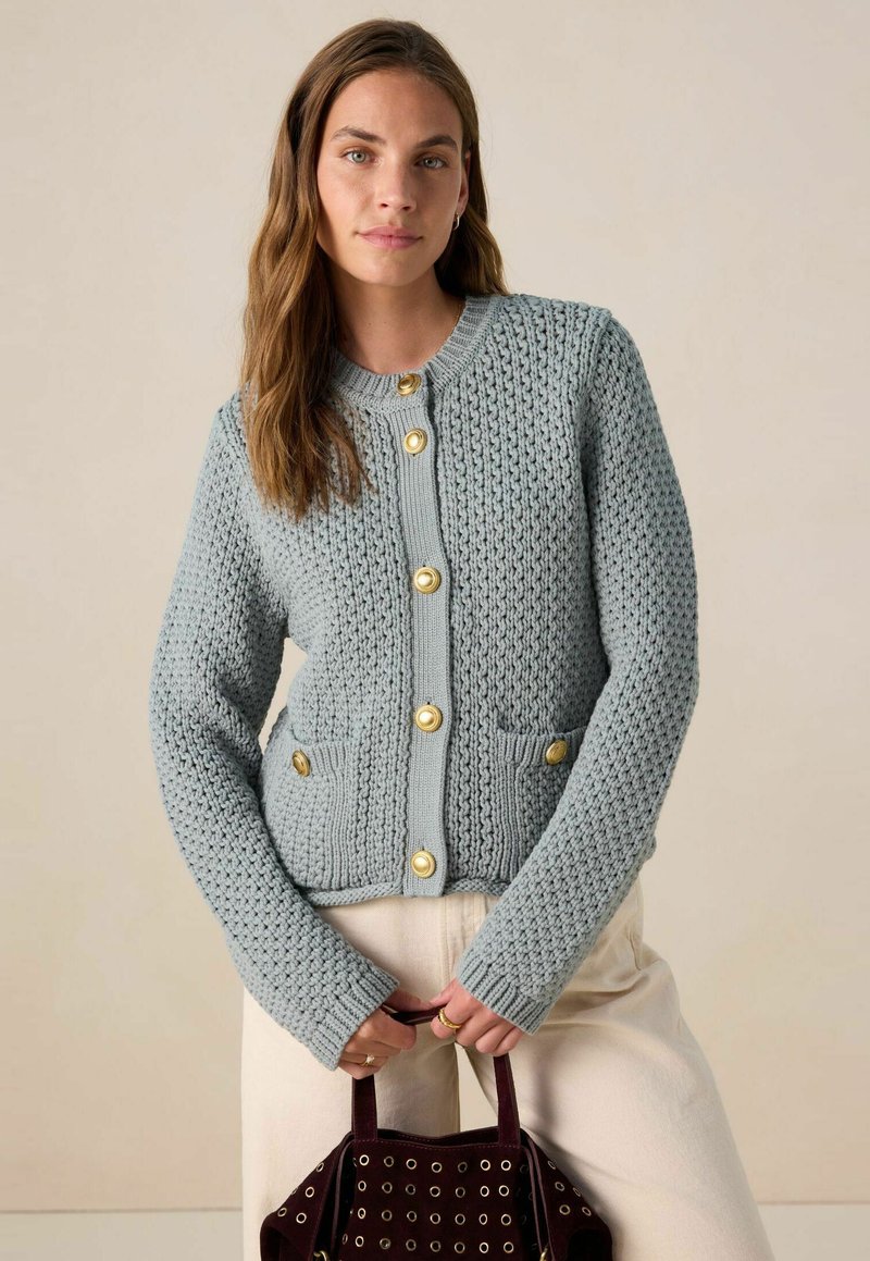Cardigan en tricot bleu clair avec un design texturé, boutons dorés et deux poches avant. Modèle tenant un sac à main noir.