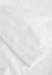 Weißer Stoff mit bestickten floralen und kreisförmigen Lochmuster, zeigt gefaltete Kanten und detaillierte Nähte.