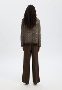 Rudas zigzaguoto rašto megztinis su per didelėmis rankovėmis, derinamas su plačiomis rudomis kelnėmis. Modelis atsisuko atgal. Matomi juodi aukštakulniai batai.