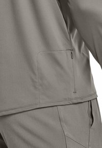 Graue Sportjacke mit glatter Textur, ausgestattet mit einer Seitentasche und kontrastierenden Nähten entlang dem Saum und den Nähten.