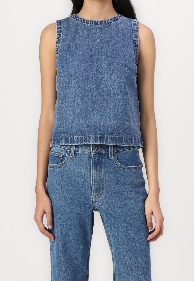 Top sin mangas de mezclilla azul, con un diseño ligeramente corto, que presenta un escote redondo y dobladillo deshilachado, combinado con jeans de tiro alto a juego.