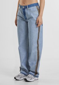 Light blue denim jeans with a dark blue waistband, featuring contrasting orange and blue side stripes. Relaxed fit with visible stitching.