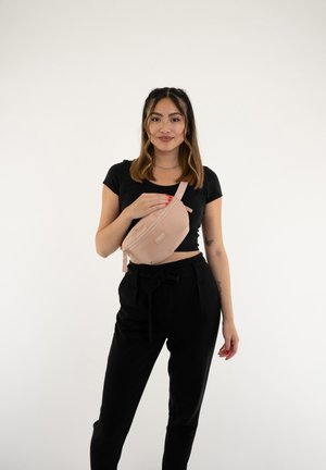 Rosa Hüfttasche aus glattem Material, mit einem Reißverschluss und einem verstellbaren Tragegurt. Das Modell trägt ein schwarzes Oberteil und Hosen.