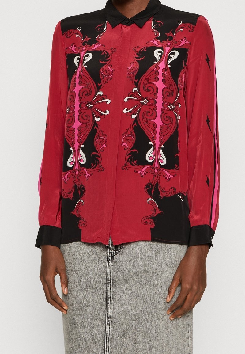 Chemise rouge à manches longues avec boutons, col et poignets noirs, ornée de motifs décoratifs symétriques noirs, roses et blancs sur le devant et d'éclairs sur les manches.