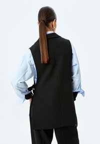 Chaleco negro sin mangas con un diseño ajustado, llevado sobre una camisa de color azul claro. Tejido suave con una forma estructurada y un detalle de abertura en la parte posterior.
