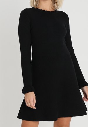 Robe en tricot noire avec un corsage ajusté, une jupe évasée, un décolleté festonné et des poignets de manches évasés. Texture douce, design minimaliste.