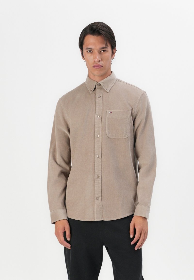 Brown button-up shirt made of textured fabric with a chest pocket, featuring a button-down collar and subtle branding on the pocket.
