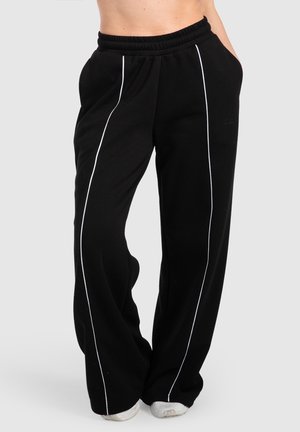Zwarte wijde broek met witte verticale bies gedragen door een persoon die staat met handen in de zakken en witte sneakers.
