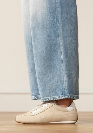 Lichtbeige suède sneakers met witte veters en hieltab, gedragen met lichtblauwe jeans met rechte pijpen op een houten vloer.