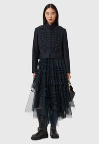 Giacca cropped blu navy con bottoni in stile militare, gonna in tulle a strati con volant in tonalità scure coordinate e stivaletti neri alla caviglia.