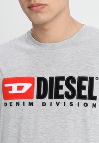 Šedé bavlněné tričko s výrazným černým a červeným logem "DIESEL DENIM DIVISION". Kolečkový výstřih a měkká textura.