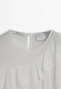 Blusa bianca con dettagli a strati di rouches, chiusura a occhietto sul retro e etichette che riportano "Massimo Dutti" e "Made in Spain". Tessuto dalla texture liscia.