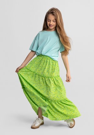 Mladé dievča s dlhými vlasmi oblečené v svetlomodrom tričku, limetkovo zelenej kvetinovej sukni a bielych teniskách, držiace a točiace sukňu.