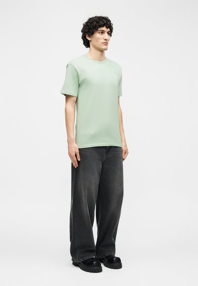 Νεαρός με σγουρά μαλλιά που φοράει ανοιχτό πράσινο μπλουζάκι, φαρδιά μαύρα τζιν και μαύρα χοντρά παπούτσια, στέκεται μπροστά σε ένα απλό λευκό φόντο.