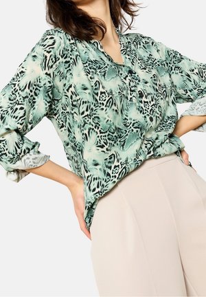 Vrouw draagt een blouse met een groen-zwarte abstracte dierenprint en opgerolde mouwen, gestoken in beige broek met hoge taille, handen in de zij.