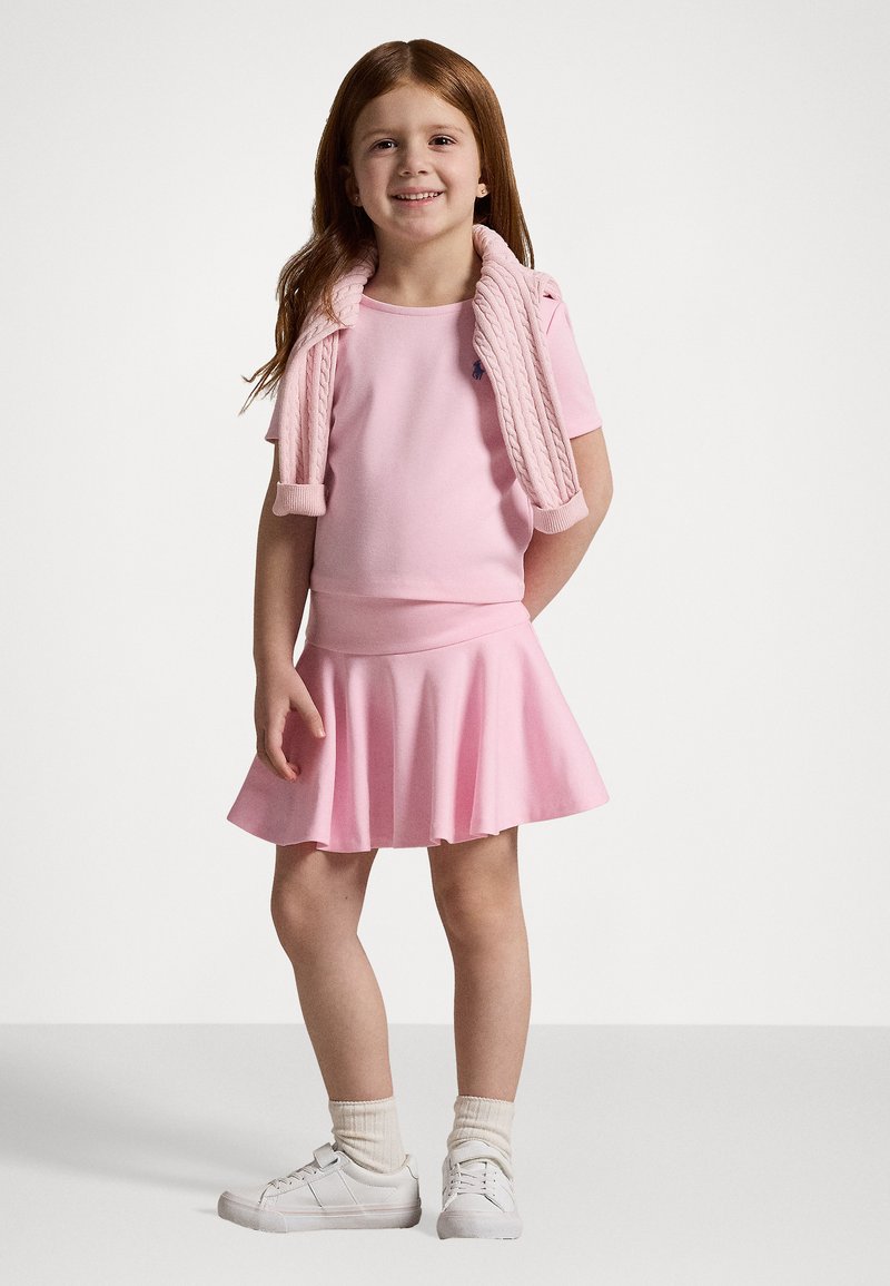 Roza obleka iz mešanice bombaža s širokim krilom, v kombinaciji s svetlo roza puloverjem z zapletenim vzorcem, ki je ovit okoli ramen, in belimi športnimi copati.