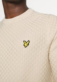 Beige Strukturpullover mit einem kleinen gelben Vogelabzeichen, das auf der linken Brust gestickt ist, getragen von einer Person mit dunkler Hautfarbe.
