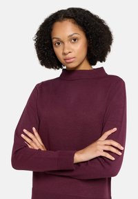 Bordowy sweter z dzianiny z wysokim kołnierzem, długimi rękawami i ściągaczami na mankietach. Tkanina jest gładka i lekko elastyczna.