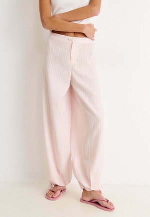 Personne portant un pantalon large rose clair et des tongs roses, debout devant un fond blanc uni, les bras croisés.