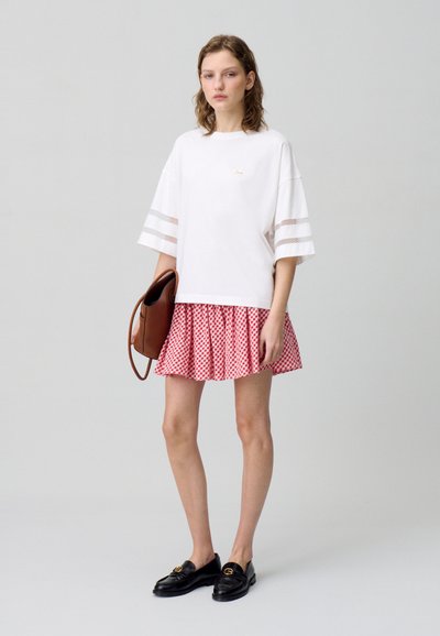 T-shirt bianco con strisce in rete, abbinato a una gonna plisettata a scacchi rossi. Una borsa marrone e mocassini neri completano l'outfit.