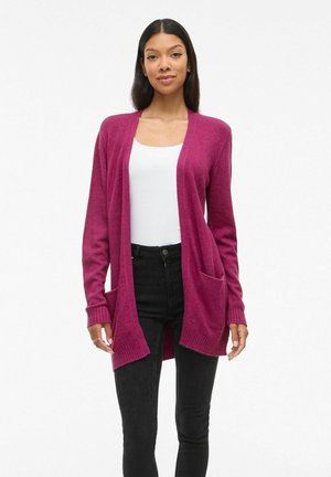Femme portant un long cardigan magenta par-dessus un haut blanc et un jean noir, debout devant un fond blanc uni.