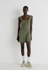 Vestido ajustado verde-azeitona com mangas curtas estilo knock e decote quadrado, com bainha evasê; usado com meias brancas e ténis bege.