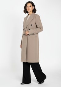 Beige dubbelknäppt kappa med slagkrage, svarta knappar och en texturerad tyg. Matchad med svarta vida byxor och spetsiga skor.