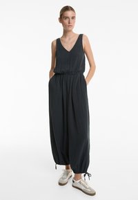 Temno siva enodelna obleka z V-izrezom, ohlapen kroj, elastičen pas in zoženi gležnji. Obuta v športne copate s bež obrobami. Gladko, teksturirano blago.