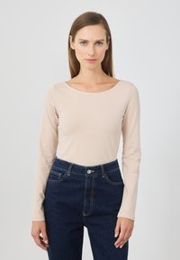 Long-sleeve top in light beige fabric with a boat neckline, paired with dark blue high-waisted jeans featuring gold stitching and front pockets.