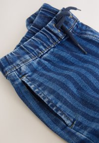 Blå denimjoggers med ett texturerat randigt mönster, elastisk midjeband och dragsko, visar gula sömmar och sidofickor.