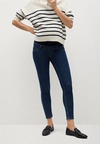 Persona vistiendo jeans ajustados de color azul oscuro, mocasines negros y un suéter blanco de manga corta con rayas horizontales negras.