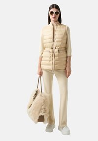 Beige gepolsterte Weste mit hohem Kragen, Kordelzug in der Taille und Reißverschlusstaschen, kombiniert mit cremefarbenen Hosen und einer flauschigen hellen Tasche. Weiße Sneakers.