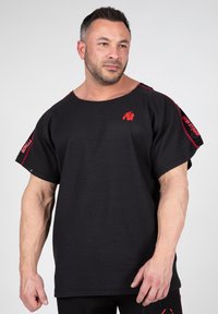 Zwart shirt met korte mouwen van een textuurstof met een brede halslijn. Rode accenten en logo-details zijn aanwezig op de schouders.