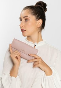 Cartera de cuero rosa claro con un acabado texturizado, que presenta un diseño de solapa simple y un nombre de marca en relieve, sostenida por una persona contra un fondo blanco.