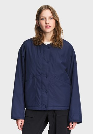 Junge Frau mit schulterlangem Haar, die eine dunkelblaue, geknöpfte Jacke und schwarze Hose vor einem schlichten hellen Hintergrund trägt.