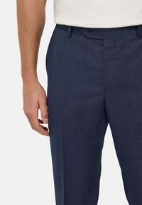 Pantalon bleu marine sur mesure en tissu texturé, avec fermeture à un bouton et devant plat. La coupe est droite avec des poches latérales.