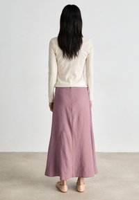 Jupe longue rayée avec des lignes verticales roses et violettes, comportant une fente au dos et un tissu doux et fluide. Associée à des chaussures de couleur claire.