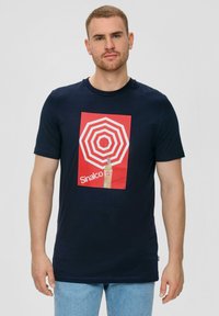 Marineblauwe katoenen T-shirt met een rode vierkante grafiek met witte concentrische cirkels en een fles, gelabeld "Sinalco." Korte mouwen.
