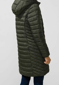 Mörkgrön, vadderad jacka med huva, med ett chevronmönster, långa ärmar och en mellanhög längd. Slätt, lätt material.