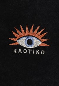 Tejido negro con un diseño bordado de un ojo con un destello solar naranja, texto "KAOTIKO" debajo en hilo blanco.