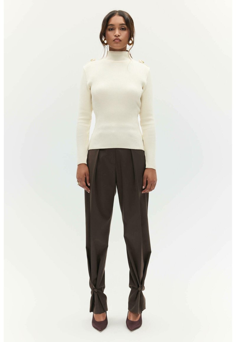 Crème ribbed coltrui met gouden schouderknopen, gecombineerd met hooglopende donkerbruine pantalon met knoopaccenten en puntige hakken.